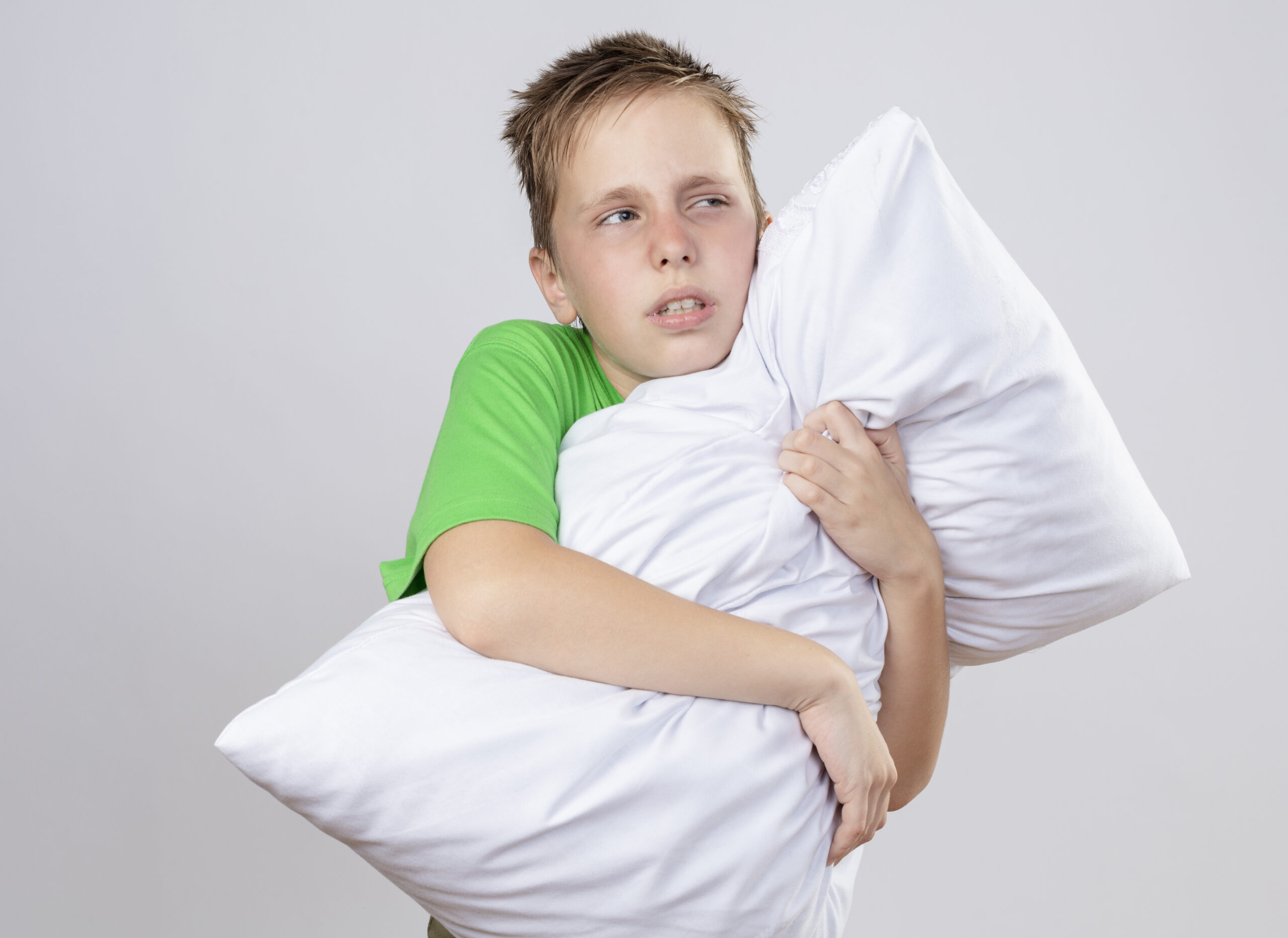 ill little boy in green t-shirt feeling unwell hugging pillow looking aside unhapy and sick standing over white background